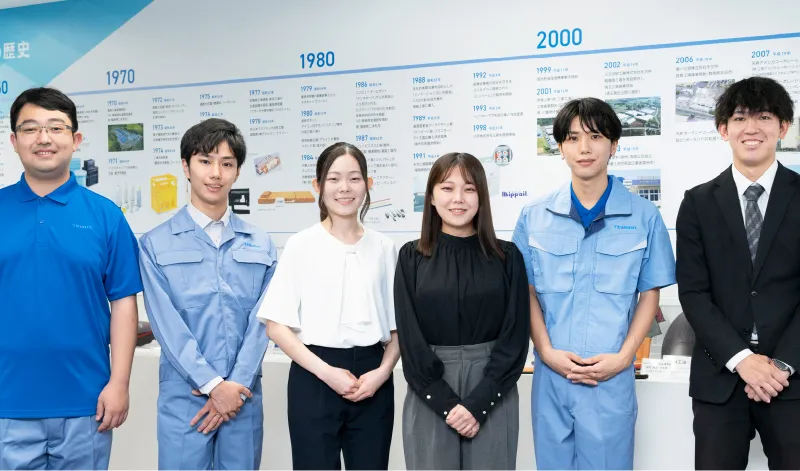 会社の歴史パネルを背景に並ぶ、スーツや作業服姿の社員たち