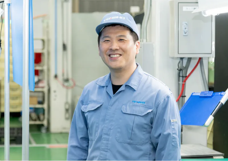 工場内で青い作業服と帽子を着用し、穏やかな表情で笑顔を見せるR.Mさん