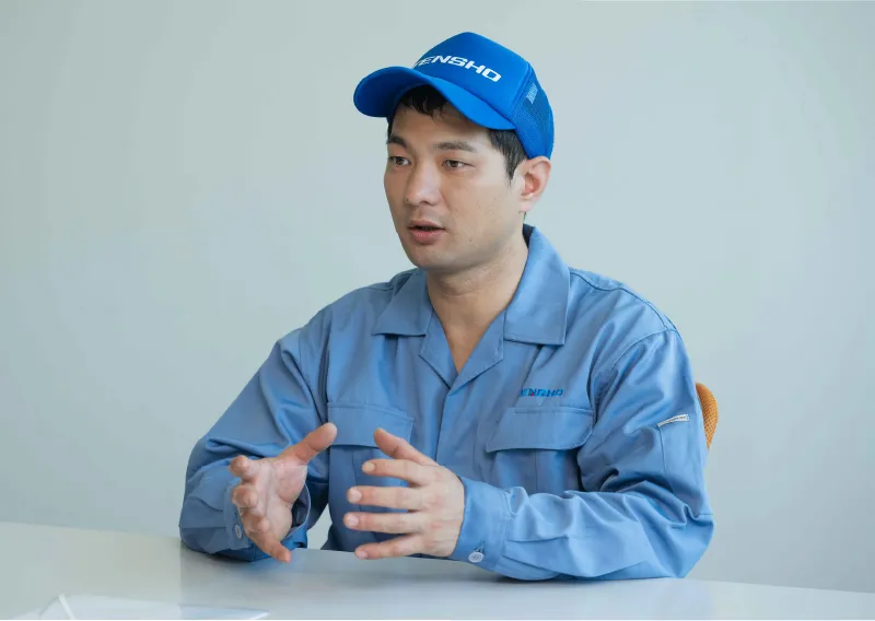 青い作業服と帽子を着用し、両手を軽く動かしながら穏やかに話すK.Aさん