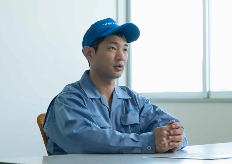 青い作業服と帽子を着用し、両手を組んで落ち着いた表情で見つめるK.Aさん