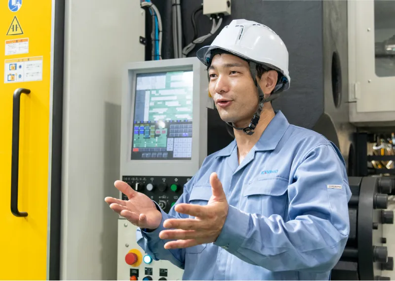 工場内で青い作業服とヘルメットを着用し、成形機の操作パネルの前で身振り手振りを交えて話すK.Aさん