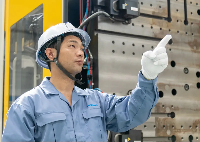 工場内で青い作業服とヘルメットを着用し、成形機の前に立って上を向いて話すK.Aさん