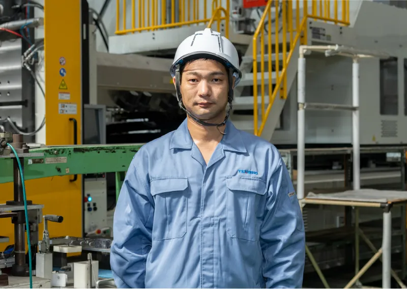 工場内で青い作業服とヘルメットを着用し、成形機の横で真剣な表情を浮かべるK.Aさん