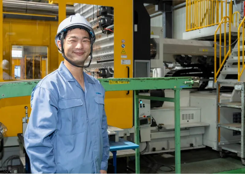工場内で青い作業服とヘルメットを着用し、大型の成形機を背にして笑顔を見せるK.Aさん