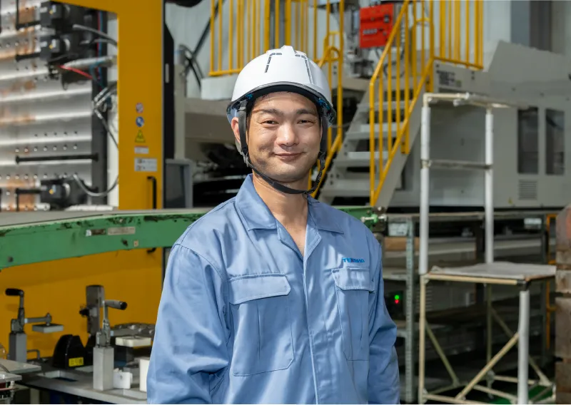 工場内で青い作業服とヘルメットを着用し、成形機の横でまっすぐ前を見据えるK.Aさん