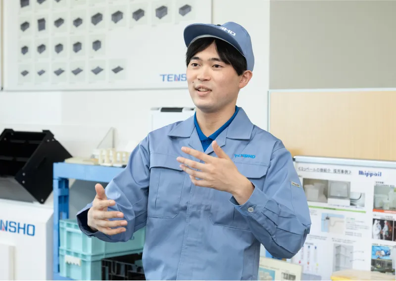 工場内で青い作業服と帽子を着用し、身振り手振りを交えて笑顔で話すK.Kさん