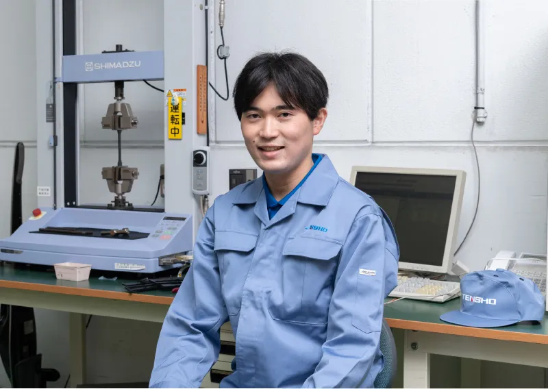 試験機の前に座り、作業服姿で穏やかな笑みを浮かべるK.Kさん