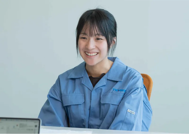 青い作業服を身にまとい、デスクに向かって優しい笑顔を見せるM.Oさん