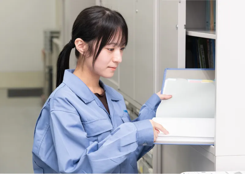 オフィス内で、棚の資料に手を添えながら横顔を見せる青い作業服姿のM.Oさん