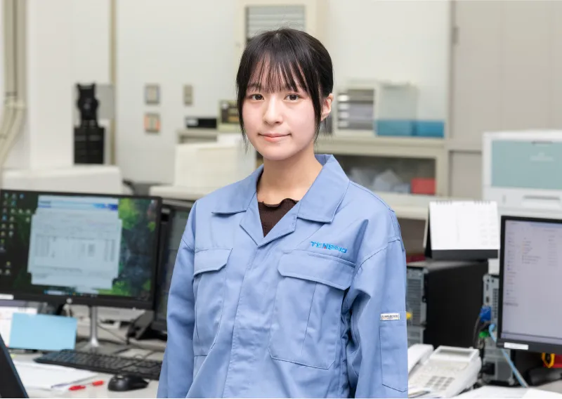 オフィス内でパソコンのモニターを背にして立ち、まっすぐ前を見つめる青い作業服姿のM.Oさん