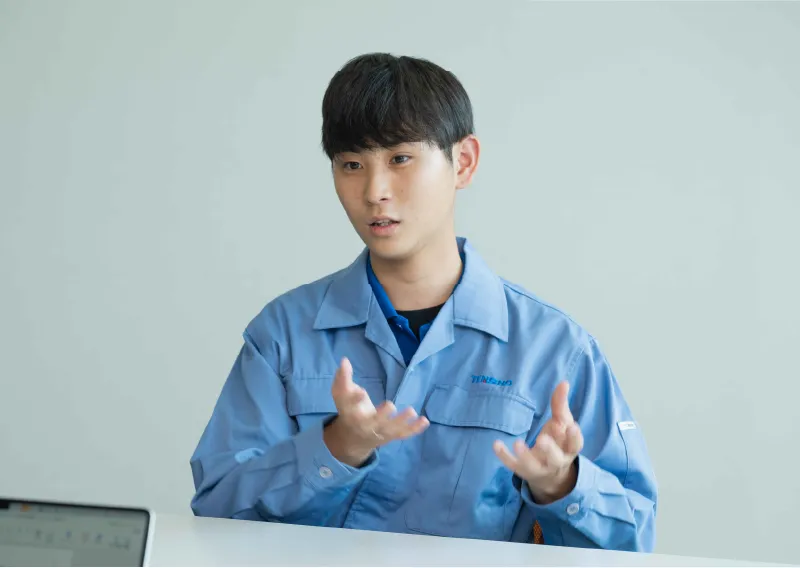 青い作業服を着用し、両手を動かしながら真剣な表情で話すO.Sさん