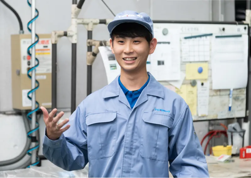 青い作業服と帽子を着用し、工場内で手を軽く差し出しながら話すO.Sさん