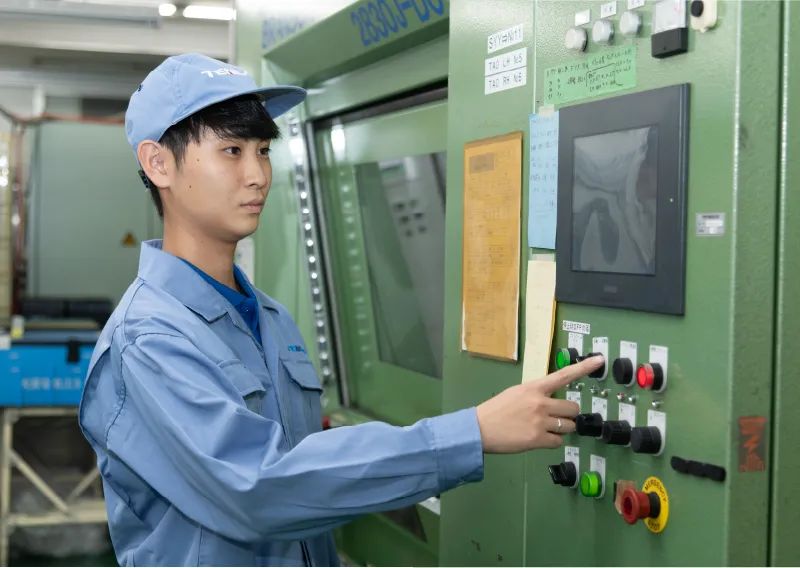 工場内で青い作業服と帽子を着用し、機械の操作パネルに向かってボタンを操作するO.Sさん