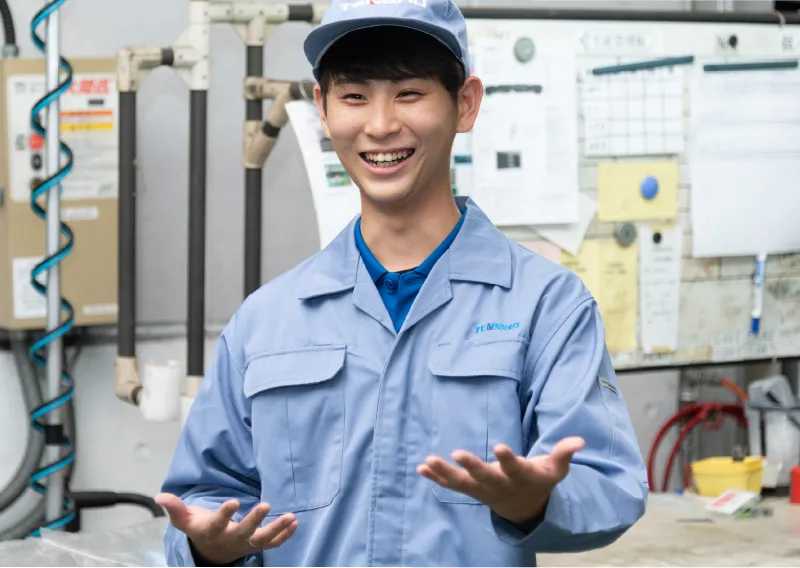 工場内で青い作業服と帽子を着用し、両手を軽く前に添えて笑顔で話すO.Sさん