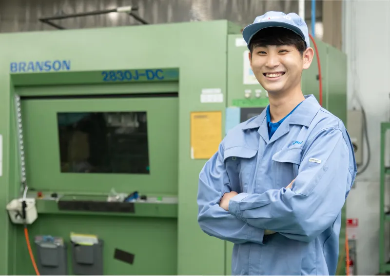工場内で青い作業服と帽子を着用し、腕を組んで笑顔を見せるO.Sさん