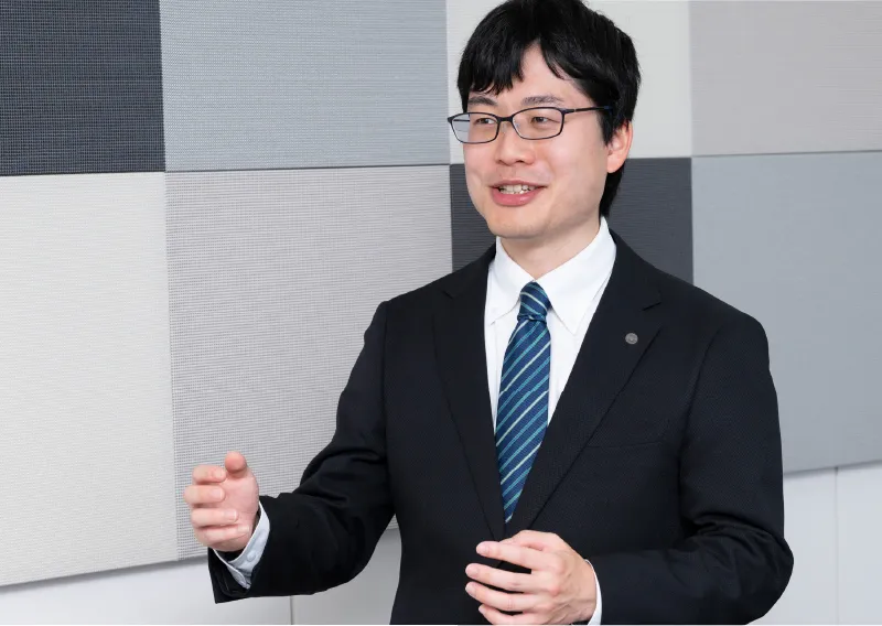 スーツ姿で身振り手振りを交えて笑顔で話す管理本部 購買部のS.Tさん