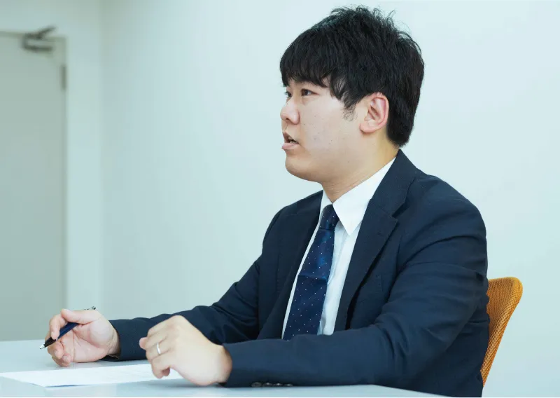 デスクに向かい、ペンを持ちながら真剣な表情で横を向くT.Fさん