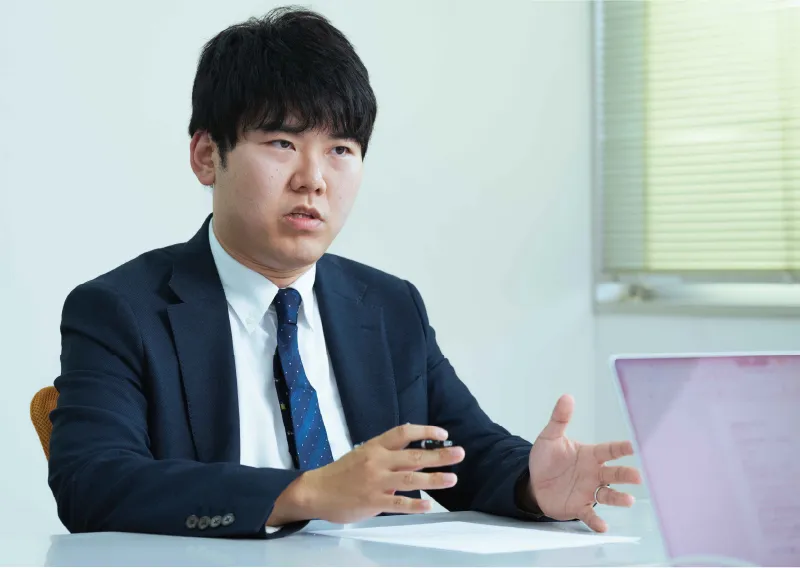スーツ姿でデスクに座り、身振り手振りを交えながら笑顔で話すT.Fさん