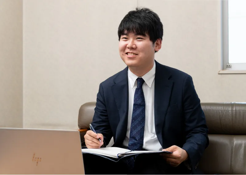 応接用ソファに座り、ノートパソコンと手帳を広げながら笑顔を見せるスーツ姿のT.Fさん