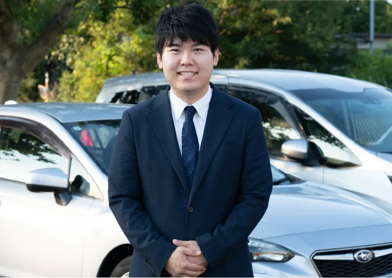 社用車の前で腕組みをしながら、穏やかな笑顔でカメラを見つめるスーツ姿のT.Fさん