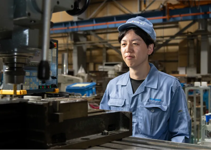 工場内で機械装置を背景に、穏やかな表情で佇む青い作業服姿のD.Hさん