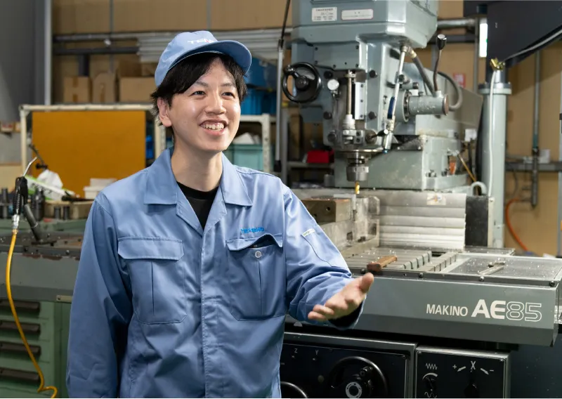 工場内の機械のそばで、笑顔を見せる青い作業服姿のD.Hさん