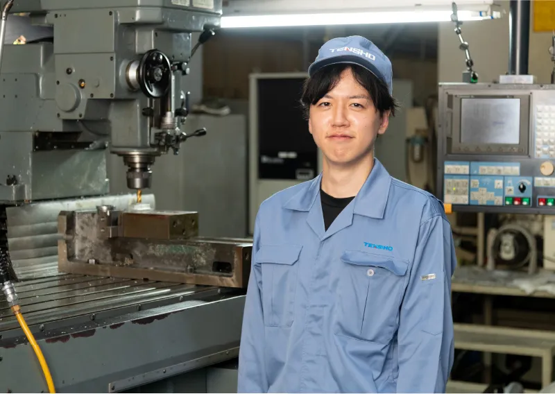 工場内で機械を背景に、落ち着いた表情で佇む青い作業服姿のD.Hさん