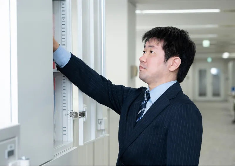 スーツ姿で棚の書類を見つめる総務人事部のT.Hさん