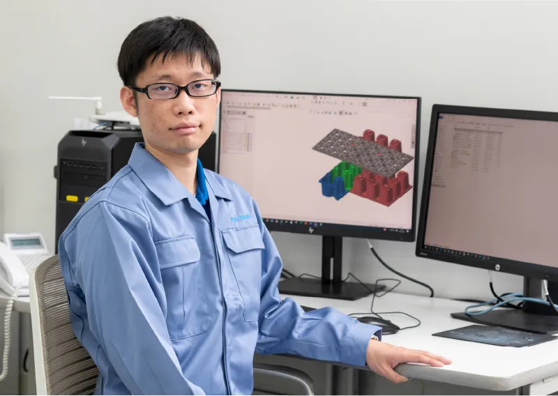 デスクで3D CADの設計画面と試作品の3Dモデルを見ながら話すT.Tさん