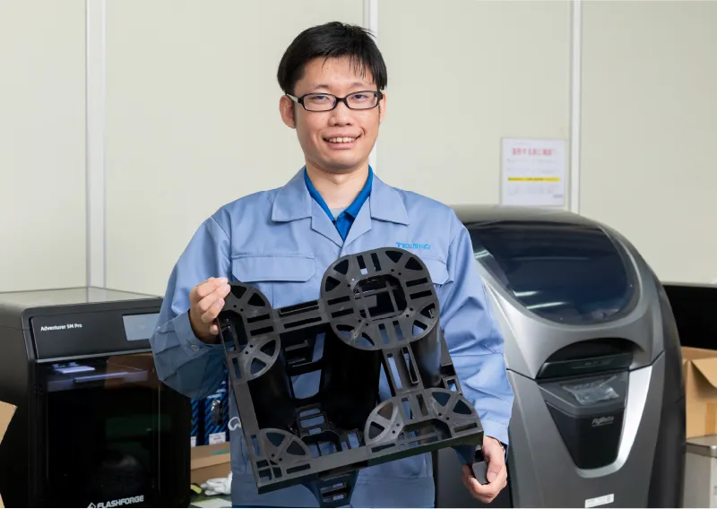 青い作業服を着用し、3Dプリンターで作製した樹脂製パーツを両手で持ちながら笑顔を見せるT.Tさん