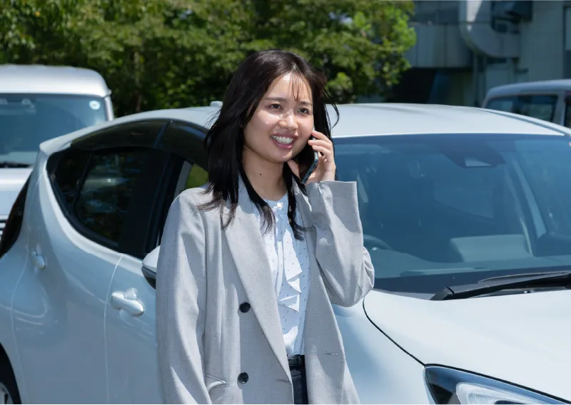 屋外の社用車を背景に、携帯電話で通話するスーツ姿のU.Oさん