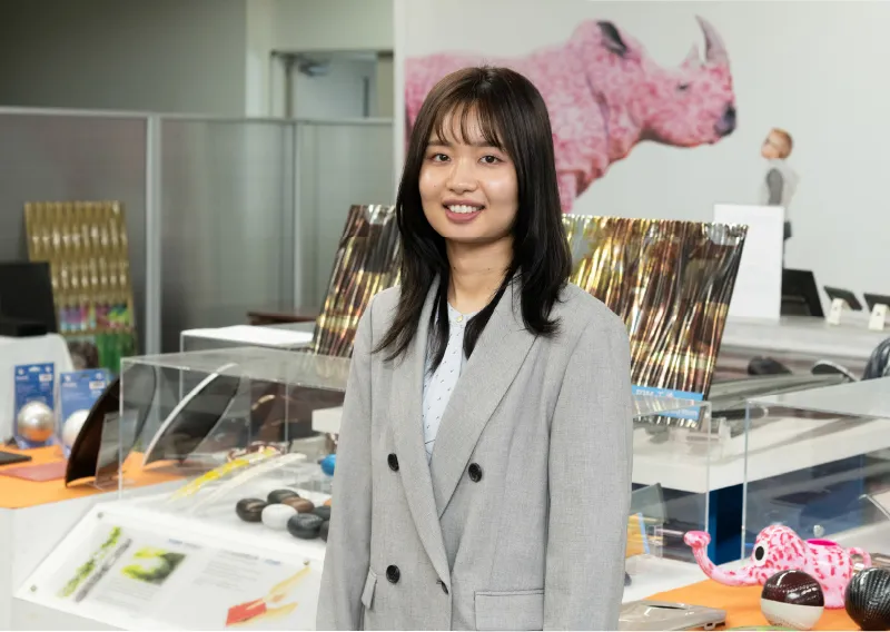 オフィスの製品を背景に、柔らかな笑顔でカメラを見つめるスーツ姿のU.Oさん