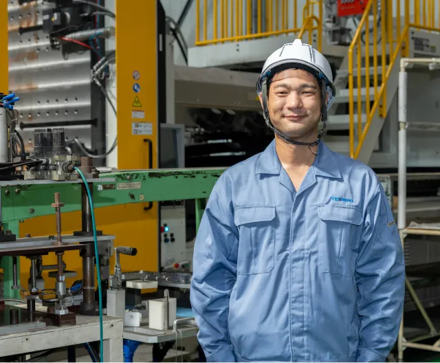 工場内の成形機を背景に立つ作業服姿の成形技能職の社員
